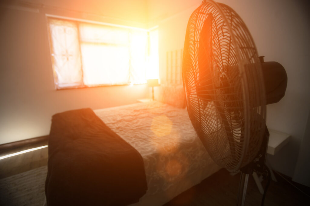 Electric fan next to bed with sunshine coming through the window.